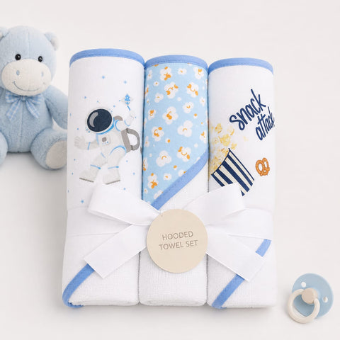 A three-pack of hooded baby towels rolled up and tied with a white ribbon, displayed on a white background with a blue teddy bear and pacifier as props. The towels feature blue trim and prints including an astronaut, daisies, and 'snack attack' popcorn.