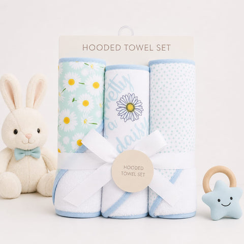 A hooded towel set displayed on a white background with a plush bunny toy on the left and a star-shaped teething ring on the right. The set includes three rolled towels: one with yellow daisy flowers on a mint green background, one with a daisy and the words 'daily daily', and one with light blue polka dots.
