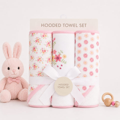 A three-pack of hooded baby towels rolled up and tied with a white ribbon, displayed on a clean white background with a plush pink bunny toy and a wooden teething ring prop.