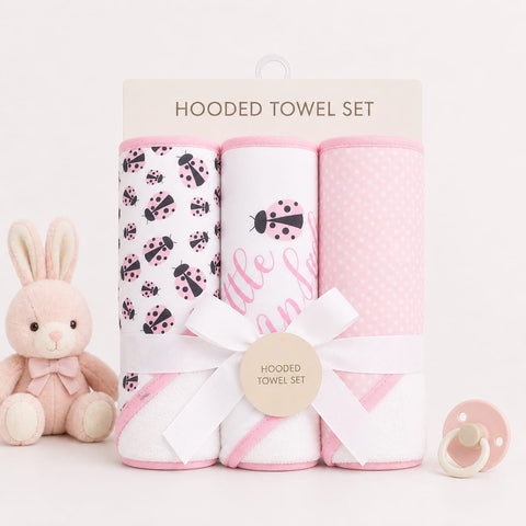 A pink and white hooded towel set in a clear plastic package with a cardboard header, tied with a white ribbon and a round tag. The set includes three towels: one with black ladybugs on a white background, one with pink script text, and one with pink polka dots.