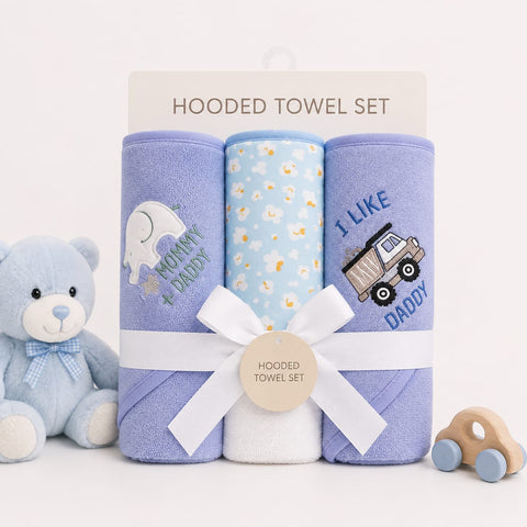 A baby hooded towel set displayed on a white background with a teddy bear on the left and a wooden toy car on the right. The set includes three rolled towels: a lavender one with a white elephant and 'KONNY PADDY' text, a light blue one with small yellow and white floral prints, and a lavender one with a dump truck and 'I LIKE DADDY' text.