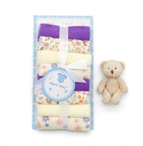 A baby gift set featuring a stack of folded baby towels in purple, cream, and floral prints, tied with a white ribbon and a round 'Alpaca + baby' tag, next to a small beige teddy bear on a white background.