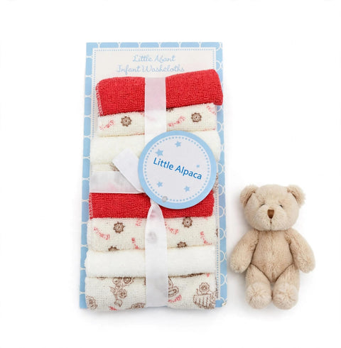 A set of Little Alpaca infant washcloths in a light blue patterned gift box, tied with a white ribbon and featuring a round blue label. A small beige teddy bear sits to the right on a clean white background.