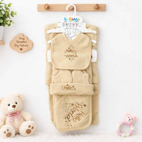 A beige baby clothing set hanging on a wooden rack against a white wall, including a bib, footmuff, and hat. The set features 'Hello World' embroidery in a gold thread.