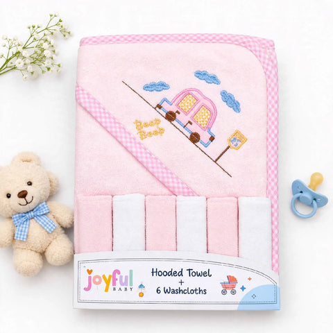 A pink hooded baby towel set with a gingham border and embroidered camper design, shown folded with six white washcloths attached. The product is photographed on a white background with a teddy bear, baby flowers, and a pacifier as styled props.