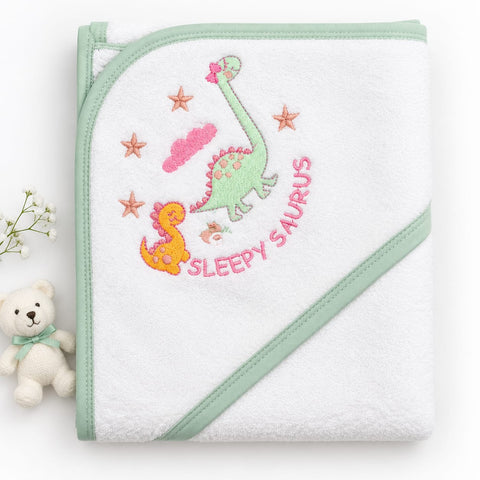 A folded white baby hooded towel with mint green trim, featuring embroidered dinosaurs, stars, a cloud, and the words 'SLEEPY SAURUS' in pink. A small white teddy bear and baby flowers sit beside it on a white background.