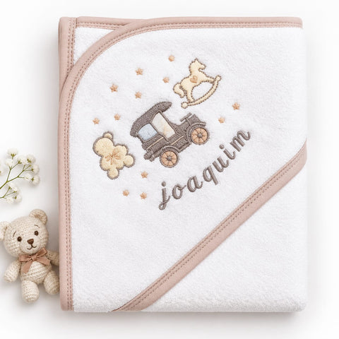 A white hooded baby towel with beige trim and embroidered motifs, including a car, rocking horse, teddy bear, stars, and the name 'joaquin' in script. A small plush teddy bear and white flowers are placed beside it on a clean white background.