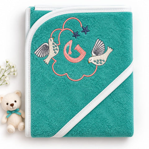 A teal baby hooded towel with white trim and embroidered birds, a pink heart, and a large letter G, shown on a white background with a small teddy bear prop.