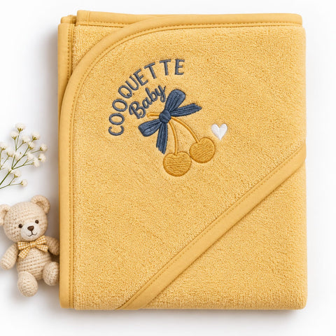 A folded mustard yellow hooded baby towel with embroidered text reading 'COQUETTE Baby' and a cherry design, styled on a white background with a small teddy bear and baby's breath flowers.