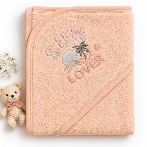 A peach-colored hooded baby towel with 'SUN LOVER' embroidered on the front, styled on a white background next to a small teddy bear and baby's breath flowers.