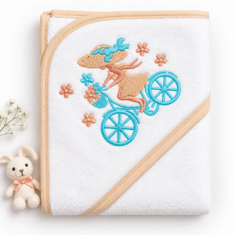 A white hooded baby towel with beige trim, featuring an embroidered design of a woman riding a blue bicycle with flowers. A small white plush bunny with a pink bow is positioned to the left of the towel on a clean white background.