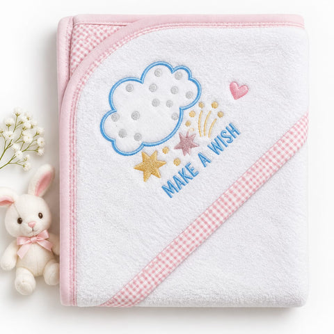 A white hooded baby towel with pink gingham trim, featuring embroidered details of a blue cloud, gold and pink stars, a pink heart, and the words 'MAKE A WISH' in blue. A small white plush bunny with a pink bow is positioned to the left of the towel on a white background.