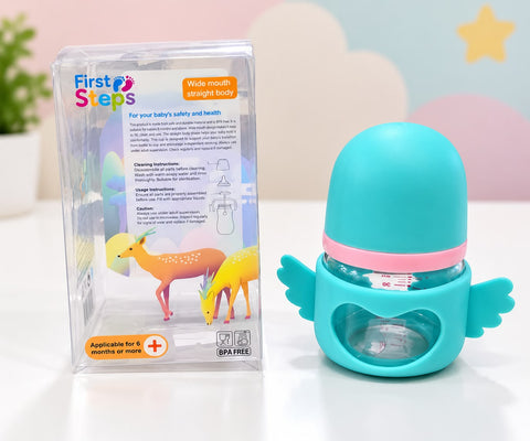 A baby bottle with a turquoise wing-shaped holder and a pink band, shown next to its clear retail packaging box on a white surface with a pastel cloud background.