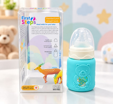 A baby bottle with a light blue silicone bear-shaped sleeve stands next to its clear plastic retail box on a white surface. The box features colorful graphics of deer and text reading 'First Steps'.