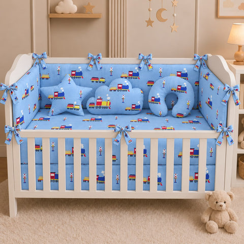 A white wooden crib in a nursery setting, styled with a light blue baby bedding set featuring a train pattern, matching crib bumper, sheet, and pillowcases, along with star and crescent moon pillows.
