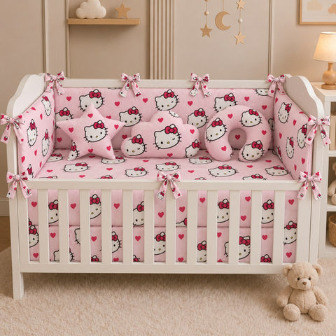 A white wooden crib in a nursery setting, fully styled with a pink and white Hello Kitty bedding set including bumper, mattress protector, sheet, and pillows. A plush teddy bear sits on the cream carpet in the foreground.