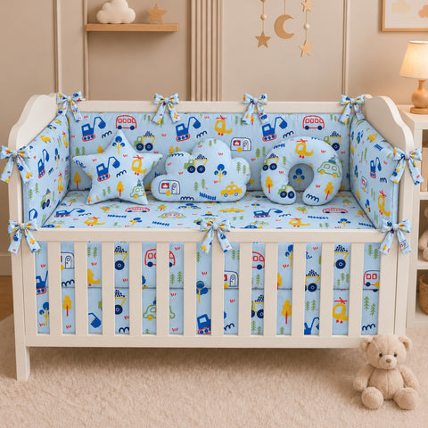 A white wooden crib in a nursery setting, styled with a light blue baby bedding set featuring a playful construction vehicle pattern in blue, yellow, and red. The set includes a fitted sheet, bumper bumpers, a crib skirt, two square pillows, and a crescent moon-shaped pillow.
