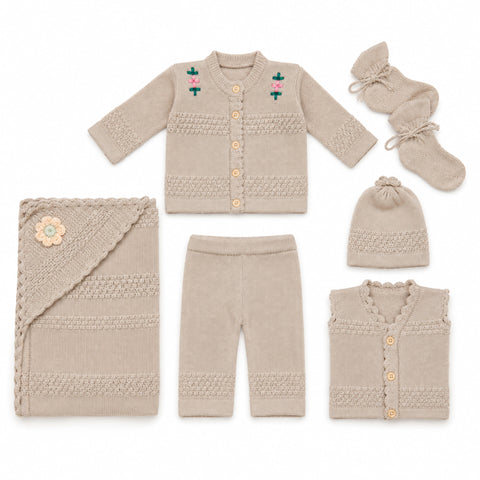 A coordinated baby knit clothing set laid out on a white background, including a cardigan with small floral embroidery, pants, a folded blanket with a flower applique, a hat, a sleeveless vest, and a pair of booties. The items are all in a warm light beige/taupe tone with small pink and green embroidered details and tan buttons.