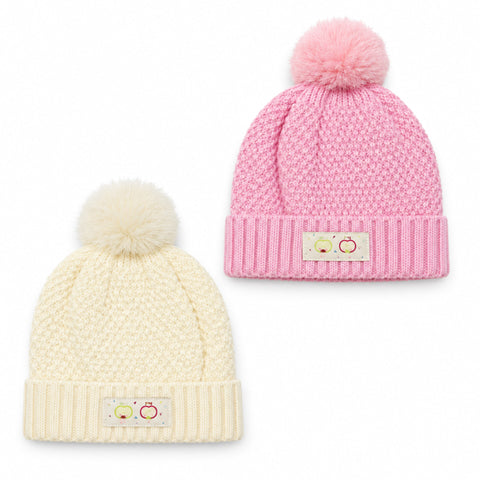 Two knit pom-pom beanies on a white background, one in cream and one in pink. Each hat has a ribbed cuff with a small decorative patch featuring two cartoon faces.