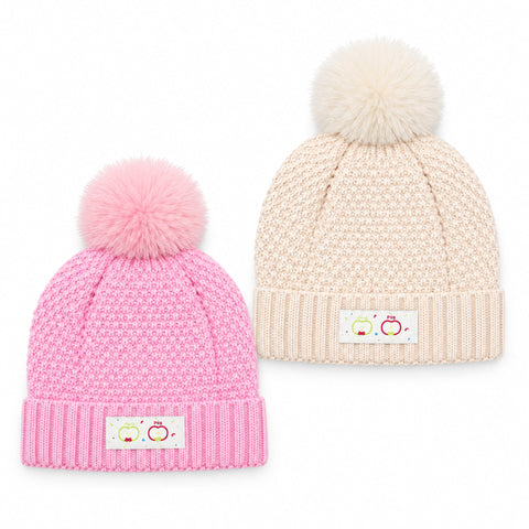 Two baby knit pom-pom beanies on a white background, one in pink and one in cream. Both have textured knit patterns, ribbed cuffs, and a small decorative patch on the front cuff.