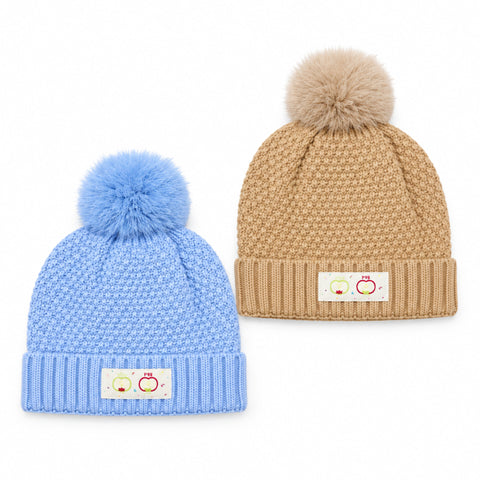 Two knit pom-pom beanies on a white background: one light blue and one tan. Each hat has a ribbed cuff with a small white patch showing three small apple illustrations.