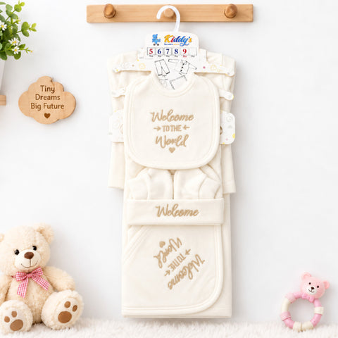 A baby clothing set hanging on a wooden hanger against a white wall, featuring a cream-colored long-sleeve top, bib, and footmuff with gold embroidered text. The set is styled with a teddy bear, a small pink toy, and a wooden sign on a white surface.