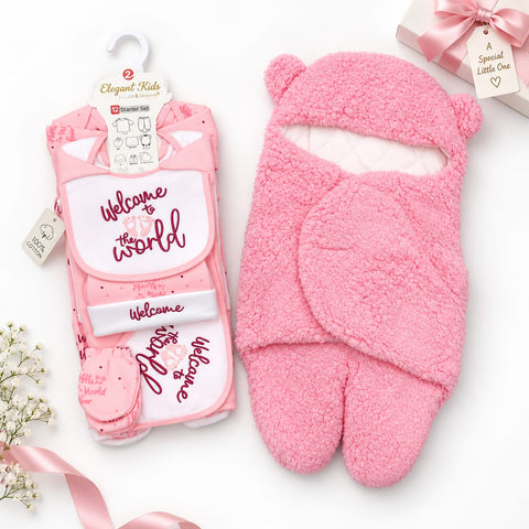 Pink baby welcome set including a plush hooded foot warmer and a matching printed bib, styled on a white background with ribbon and flower props. The printed bib reads 'Welcome to the world' and the foot warmer has bear ears.