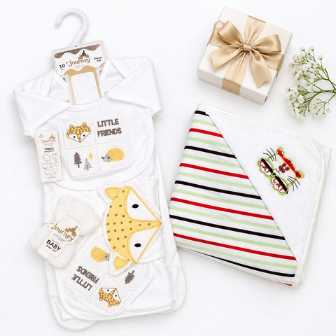 A baby gift set arranged on a white background, including a white hooded baby outfit with embroidered animal faces and text, a striped white baby blanket with a tiger face patch, a small white gift box tied with a beige ribbon, and a sprig of white flowers.