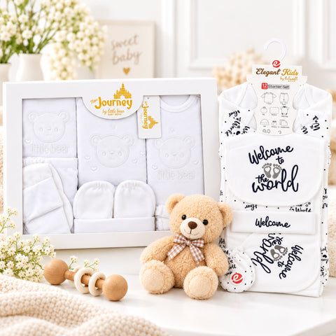 Baby gift set featuring a white boxed clothing set with a teddy bear face print, a matching white bib, a white footmuff, and a small plush teddy bear with a bow tie, styled on a light surface with flowers and a wooden rattle.