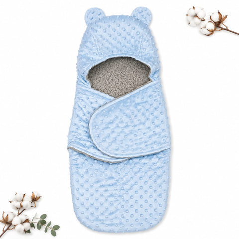 A light blue baby swaddle wrap with a textured minky dot pattern, featuring bear ears on the hood and a soft gray fleece lining. The swaddle is displayed flat on a white background with decorative cotton bolls in the corners.