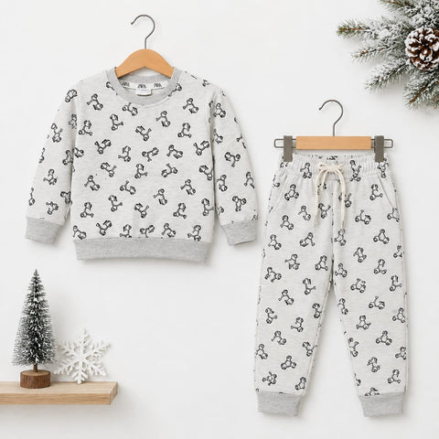 A children's two-piece pajama set hanging on wooden hangers against a white wall. The set includes a long-sleeve sweatshirt and matching jogger pants in a light gray fabric with a black repeating graphic pattern, styled with small holiday props including a miniature tree, snowflake, and pine branch.