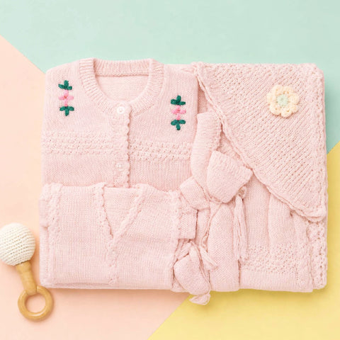 A folded light pink knitted baby outfit set on a pastel geometric background, including a cardigan-style top with embroidered flowers, matching bottoms, a knitted wrap or cape with a flower applique, and a small white-and-wood rattle nearby.
