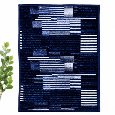 A rectangular quilted textile with an abstract geometric pattern in navy blue, white, and lighter blue stripes, displayed flat against a white background with a small green leafy branch on the left.