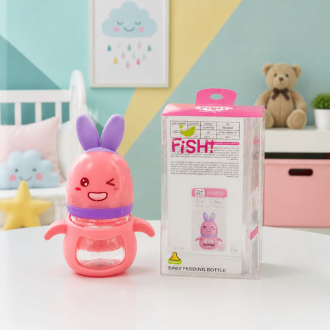 A pink bunny-shaped baby feeding bottle with purple ears and a purple collar sits on a white table next to its retail packaging box. The background shows a colorful children's room setting with a teddy bear, pillows, and a framed cloud print.