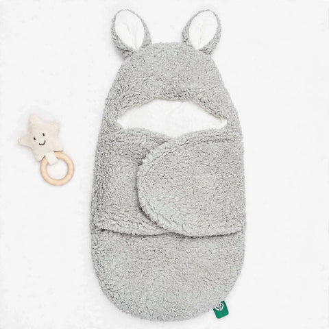 A light gray plush baby swaddle blanket with bunny ears and a matching footed cowl, shown on a white background next to a small star-shaped wooden teething toy.