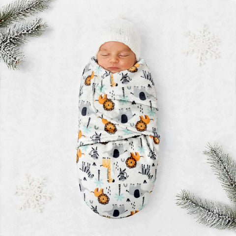 A sleeping newborn baby swaddled in a white blanket with a colorful safari animal print, wrapped in a soft white knit hat, laid flat on a white textured surface with snowy decorative props.