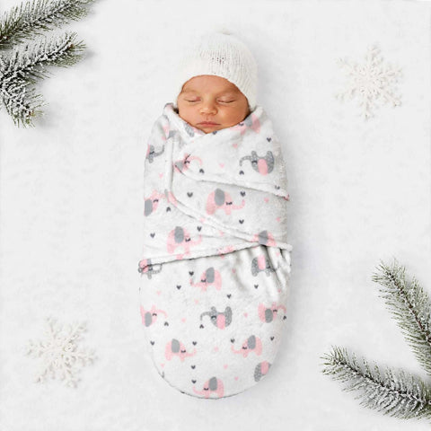 A sleeping newborn baby swaddled in a soft white blanket with a pink and gray elephant print, wearing a white knit hat, laid on a white background with decorative pine branches and snowflakes.