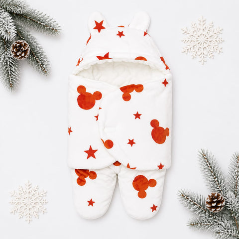 A baby footed swaddle blanket with a hood and ear details, featuring a white background with red Mickey Mouse head silhouettes and red stars. The swaddle is styled in a flat-lay with winter-themed props including pine branches, pinecones, and white snowflakes on a light gray background.
