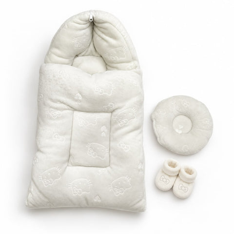 A baby sleep bag set in warm off-white/cream, featuring a quilted hooded sleeping bag with a central pocket and subtle Hello Kitty face patterns, accompanied by a matching round cushion and two small booties on a clean white background.