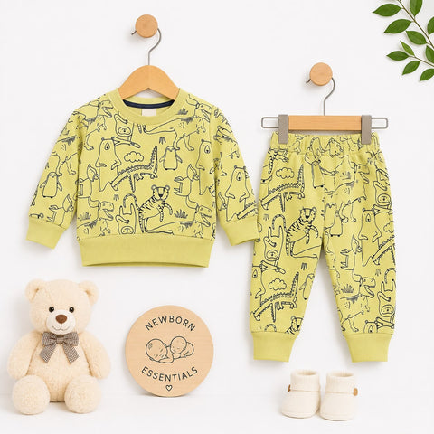 A baby clothing set displayed on wooden hangers against a white background, including a long-sleeve sweatshirt and matching jogger pants in a pale yellow-green color with black animal line drawings. A teddy bear, a round 'Newborn Essentials' card, and cream booties are arranged around the set.