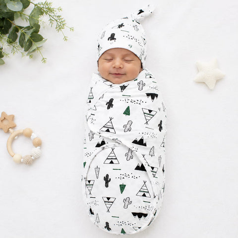 A sleeping newborn baby swaddled in a white muslin swaddle blanket with a black and green geometric teepee and cactus pattern, wearing a matching beanie. The baby is photographed on a white fabric background with baby props including a wooden star teether, a green eucalyptus sprig, and a white star toy.