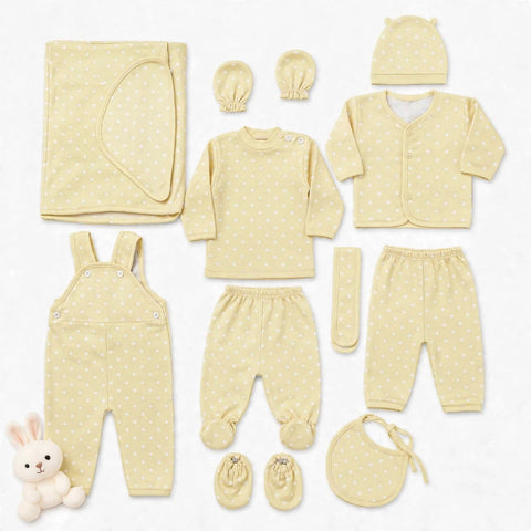 A coordinated baby clothing set laid out on a white background, including a long-sleeve top, footed pants, overalls, cardigan, hat, booties, bib, mittens, and a folded blanket. The set is a soft pale yellow with small white polka dots, and a small plush bunny toy appears in the lower left corner.