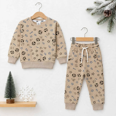A children's two-piece outfit set consisting of a beige long-sleeve sweatshirt and matching jogger pants, both decorated with an all-over print of black soccer balls, blue stars, and brown scribbles. The sweatshirt is shown on a wooden hanger on the left, and the pants are shown on a metal hanger on the right, set against a white background with holiday-themed decorative props.