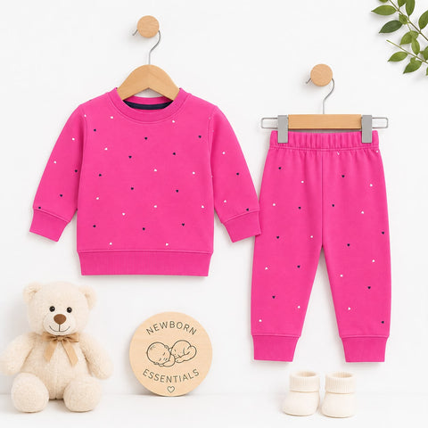 A baby clothing set displayed on wooden hangers against a white background, featuring a bright pink long-sleeve sweatshirt and matching jogger pants with small scattered polka dots. A cream teddy bear sits at lower left, a round wooden sign reading 'Newborn Essentials' is centered, and small cream booties appear at lower right.