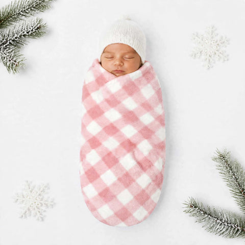 A sleeping newborn baby swaddled in a pink and white gingham blanket wrap, wearing a white knit beanie, laid flat on a white background with decorative snowy snowflakes and pine branches in the corners.