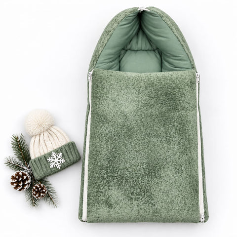 A sage green fleece baby footmuff with white zipper trim, shown next to a matching knit pom-pom beanie and pinecones on a white background.