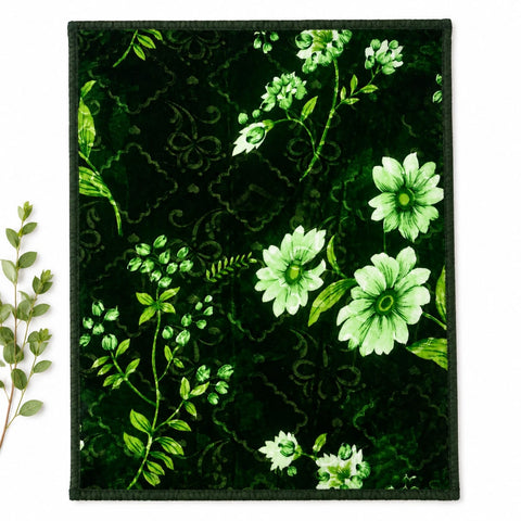 A square fabric item, likely a small blanket, throw, or mat, featuring a dark green velvet-like background with a lighter green damask pattern and white and light green floral motifs. The item is displayed flat against a white background with a small decorative leafy sprig on the left side.