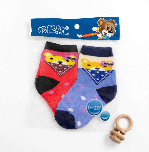 A pair of baby ankle socks in red and blue, featuring cartoon bear faces with bandanas, shown in their blue retail packaging with a wooden teether toy on a white background.