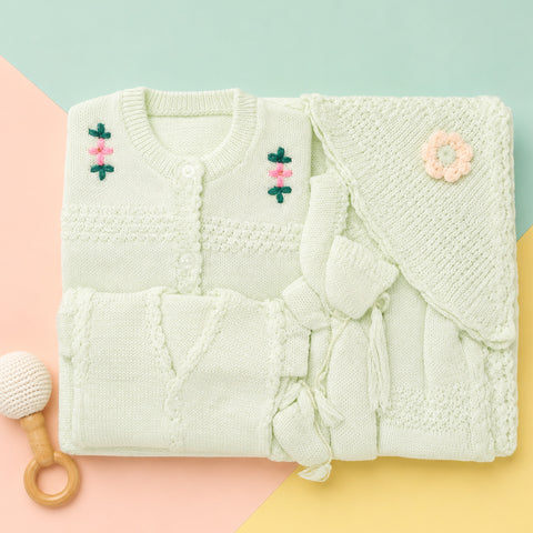 A folded pale mint green knitted baby outfit set on a pastel geometric background, including a long-sleeve cardigan with embroidered flowers, a matching hat, and a small rattle.