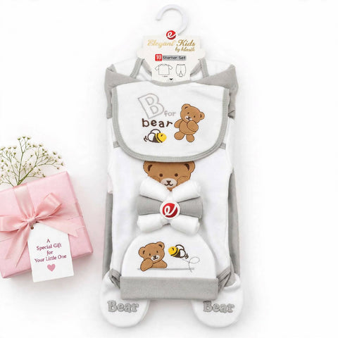 A baby footie sleeper set by Elegance Kids by Heart, featuring a white and gray bib-style top with a brown teddy bear graphic, a gray bow, and matching footie socks. The set is displayed on a white background next to a pink gift box with a baby's name tag.